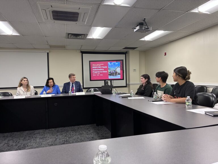 Secretary Lee, Deputy Secretary Michael Lore, and Rosanna Mantova hear the Youth Ambassadors' report on the trip Three officials seated at a conference table with name cards face three students across from them, with a presentation slide on a wall screen