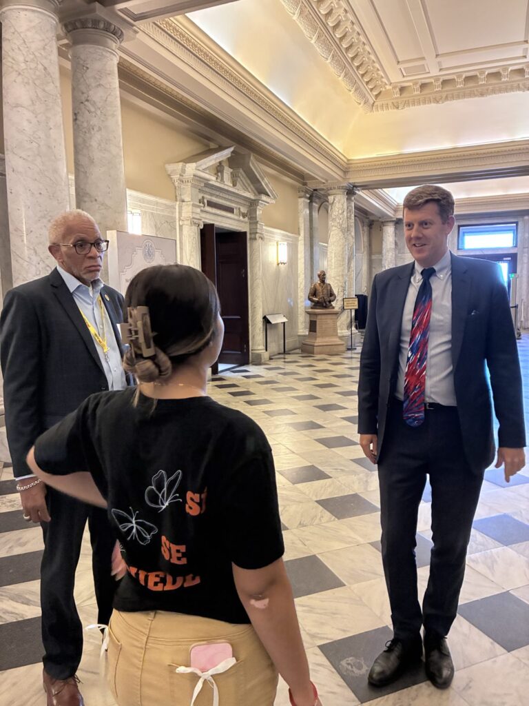A Youth Ambassador talks with Deputy Secretary Lore in the Maryland State House A student talks with two men in suits in a marble hallway with columns outside the House Chamber
