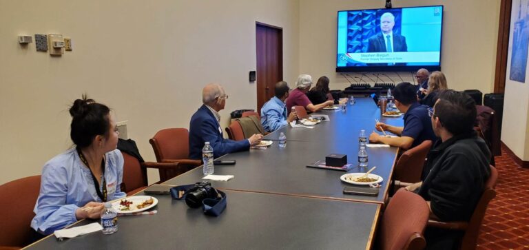 Attendees seated along a conference table at Baltimore City Hall watching a webcast of Stephen Biegun, former Deputy Secretary of State, on a wall screen